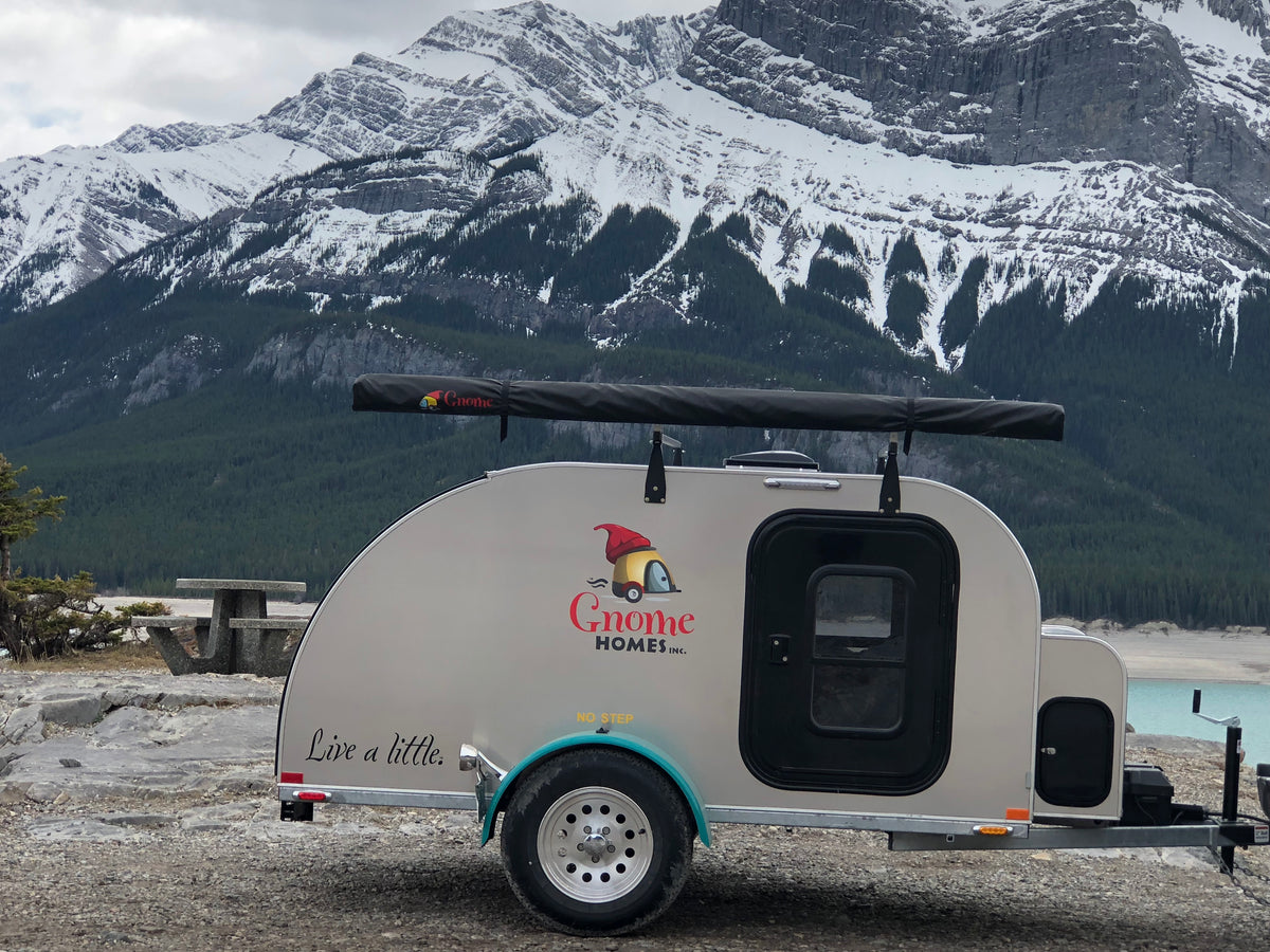 A Cruiser trailer parked outdoors in front of a snowy mountain landscape. The trailer is silver with turquoise trim, a awning, and visible branding that reads 'Gnome Homes' with a gnome logo.
