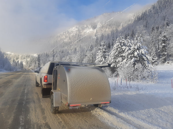 A small covered trailer attached to a white pickup truck on a snowy mountain road, with snow-covered trees and a misty blue sky in the background.